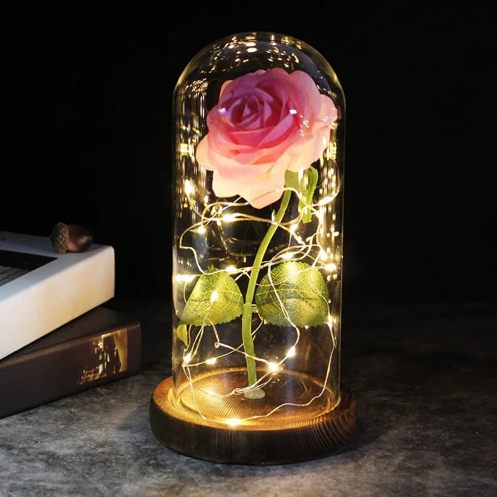 Artificial rose under a clear dome with LED string lights around the stem, on a round wooden base - pink