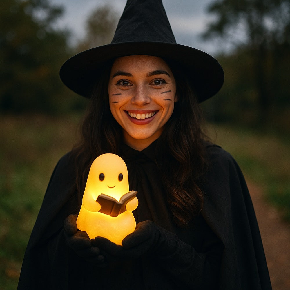 White ghost lamp with black eyes and pink cheeks holding an open brown book, glowing warm yellow