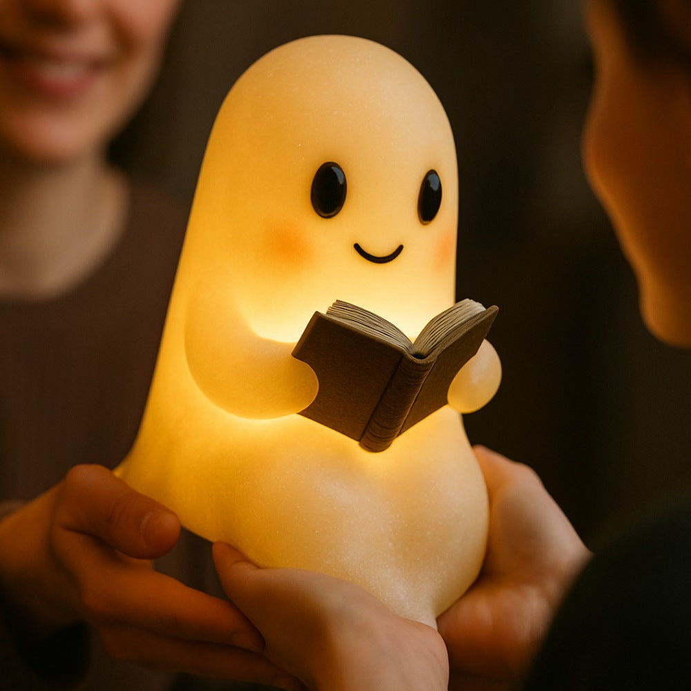 White ghost lamp with black eyes and pink cheeks holding an open brown book, glowing warm yellow