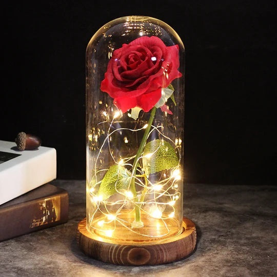 Artificial rose under a clear dome with LED string lights around the stem, on a round wooden base - red