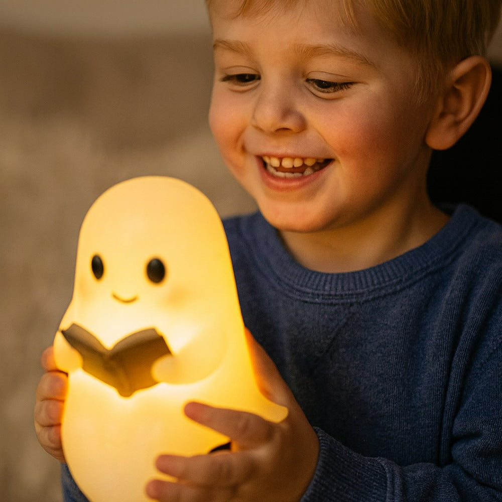 White ghost lamp with black eyes and pink cheeks holding an open brown book, glowing warm yellow