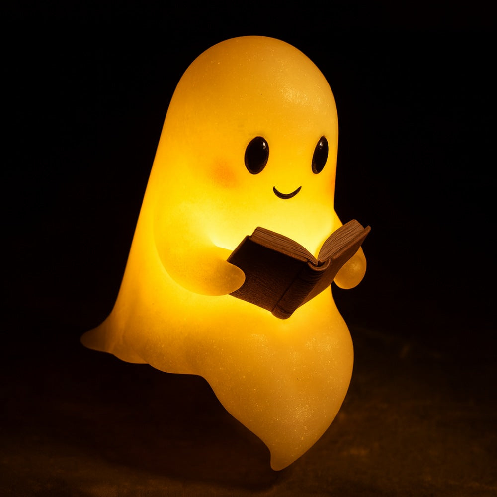 White ghost lamp with black eyes and pink cheeks holding an open brown book, glowing warm yellow